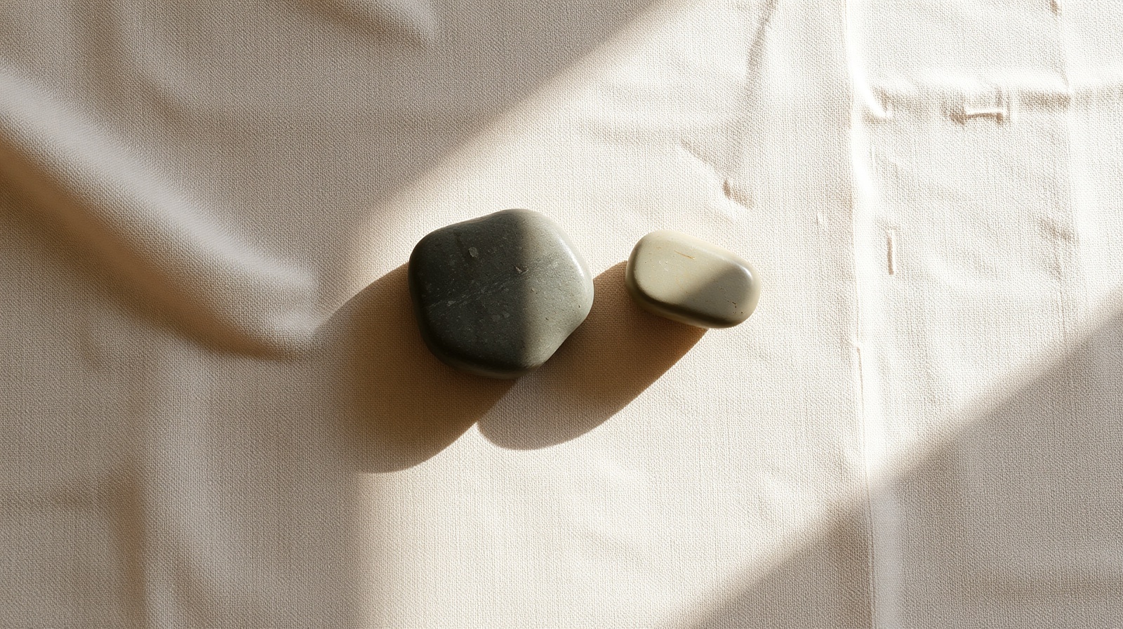 Two smooth river stones balanced beside each other on warm linen, directional morning light, neutral palette with a single persimmon thread.